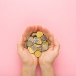 Opened young adult woman palms with euro coins on light pink table background. Pastel color. Point of view shot. Closeup. Top down view.